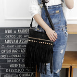 A woman wearing a white top and denim overalls holds a fringed black bag adorned with studs, posing in a stylish interior.