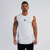 A muscular man stands in a sleeveless white shirt, showcasing his tattoos against a neutral backdrop.