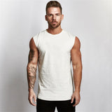 A muscular man stands confidently in a sleeveless white shirt, showcasing detailed arm tattoos against a plain background.