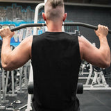 A muscular man in a sleeveless black shirt performs a pull-up, lifting himself on a bar in a gym setting with equipment in the background.