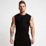A muscular man stands confidently in a black sleeveless shirt, showcasing his tattoos and strong physique against a plain background.