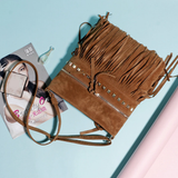 A stylish brown suede saddle bag with fringe detailing and stud accents is placed beside a magazine and a glass dropper on a pastel surface.
