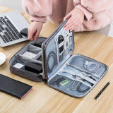 A person organizes electronic accessories in a gray travel case, surrounded by a laptop, phone, and pen on a wooden table.