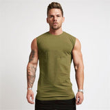 A muscular man stands confidently in a sleeveless olive green shirt, showcasing his tattoos and strong physique against a plain background.
